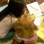 A young girl with long black hair, wearing a pink and white striped shirt, "reads" books with a Golden Retriever pup.