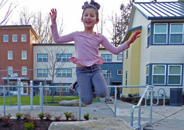 Young girl wearing a pink shirt and grey pants jumps off a boulder on a playground. Behind her is housing.