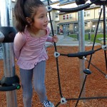 Young girl wearing a pink shirt and grey pants plays near a jungle gym.