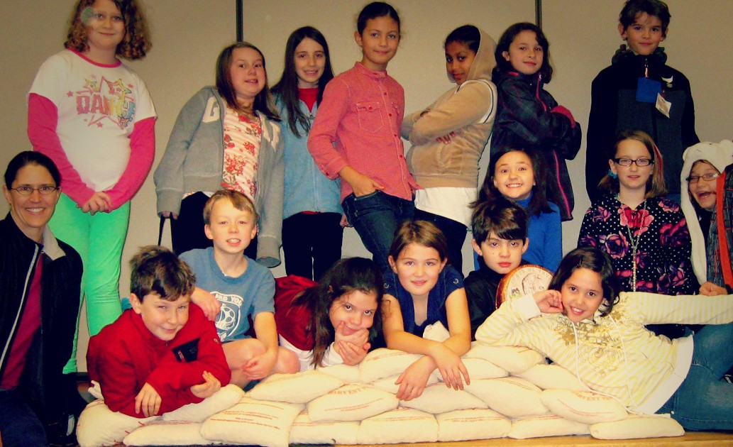 A group of 16 elementary school students and one teacher pose with a mound of sacks filled with coins.