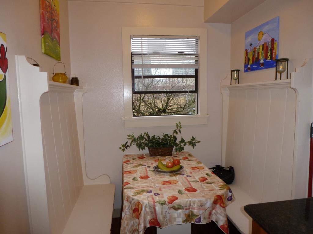 Picture of a painted white breakfast nook with a table and cheerful tablecloth, potted plant, window, and colorful art on the walls.