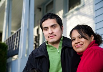 Latino man with mustache in a green shirt and jacket and Latina woman in red top smile in front of an apartment building