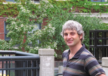 White man with grey hair wearing a striped polo shirt, standing outside in front of a brick building and a green leafy tree