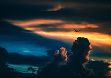 Airplane flying through a dramatic cloudy, sunset sky over mountains and an aquamarine body of water