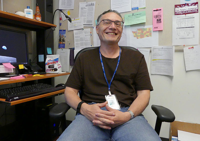 Man wearing glasses, a black shirt, jeans, and a lanyard with nametag sits in an office chair in a work cubicle.