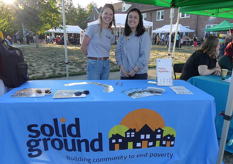 Two young women in grey T-shirts stand behind a table with a blue tablecloth and the Solid Ground logo on it