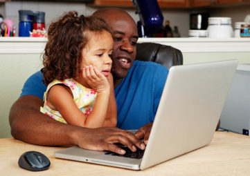 A father and his small daughter look at a laptop together at their kitchen table
