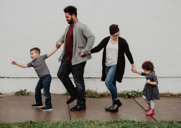 A multi-racial family of 4 holds hands & walks down a wet sidewalk in rainboots