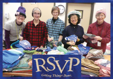 Five women wearing knitted hats, standing in front of a table full of knitted items, with a blue RSVP tablecloth logo in front of it.