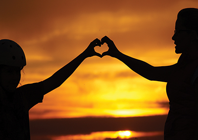 Silhouette of a mother & daughter holding their hands up to form a heart in front of a brilliant orange sunset