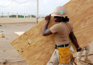 Black woman construction worker in hard hat and sunglasses wearing khaki clothes carries a large piece of plywood across a sandy lot