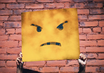 Person holding a yellow square sign with an angry face painted on.