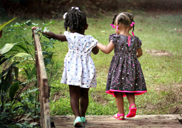 Two young girls holding hands and walking across a brown bridge.