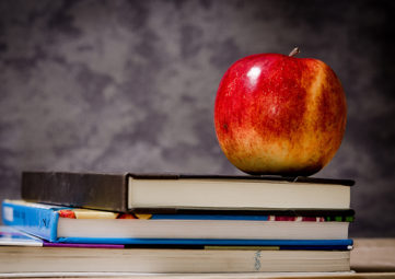 Stack of 3 textbooks on a table withe a red apple sitting on top, in front of a grey background