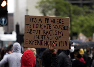 Group of protesters viewed from the back, one holding a sign that reads 'It's a privilege to educate yourself about racism instead of experiencing it!!!'