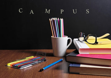Brown table with colored pencils lying on it and in a white mug. Stack of 3 hard-bound books with eyeglasses on them. Blackboard in the background with the word CAMPUS on it.