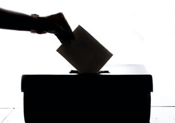 Black & white silhouette of a woman's braceleted hand dropping a ballot into a voting box.