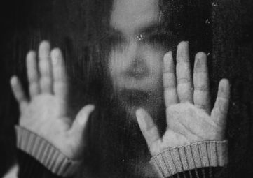 Black & white photo of a woman staring mournfully out of a rain-streaked window with her hands pressed against the glass.