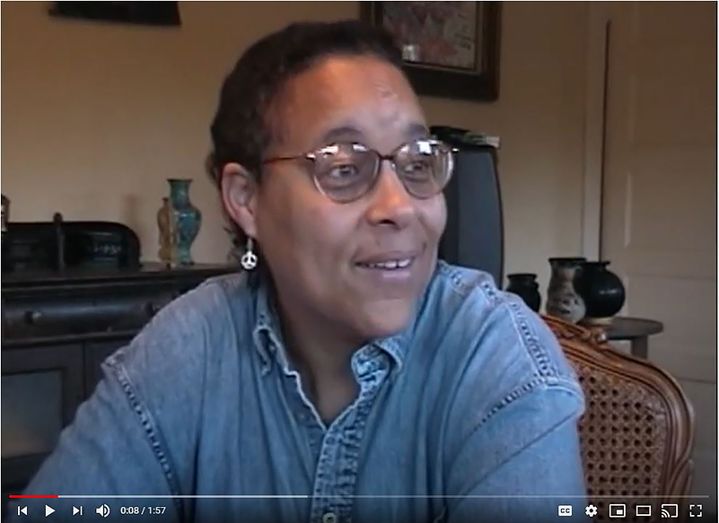Screenshot of a YouTube video of a woman being interviewed. She sits in a wicker chair and is wearing a blue denim shirt, peace symbol earrings, and wire-framed glasses.