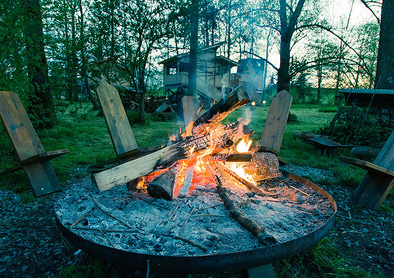 Bright orange campfire surrounded by 5 wooden chairs with the cabin in the background.