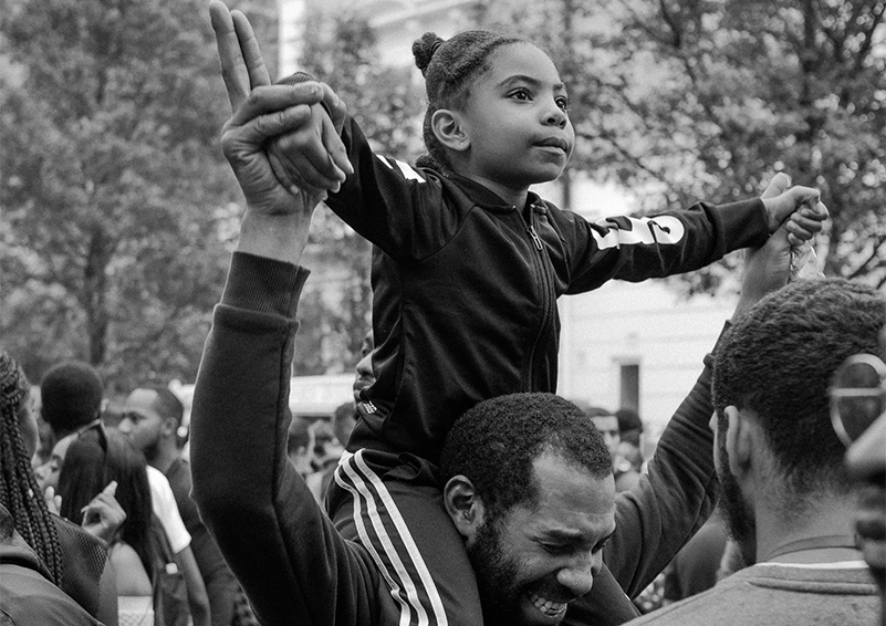 Grayscale photo of a young girl sitting on a man's shoulders with both their hands in the air at an outdoor group gathering