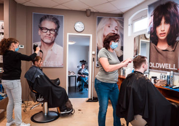 A salon with two hairdressers with masks on styling hair more than 6 feet apart, with larger-than-life sized posters of people on the walls