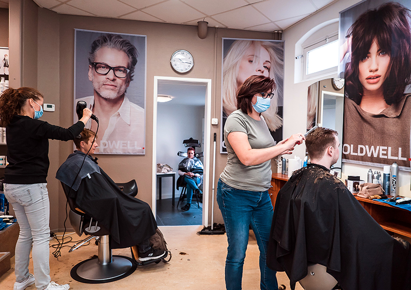 A salon with two hairdressers with masks on styling hair more than 6 feet apart, with larger-than-life sized posters of people on the walls