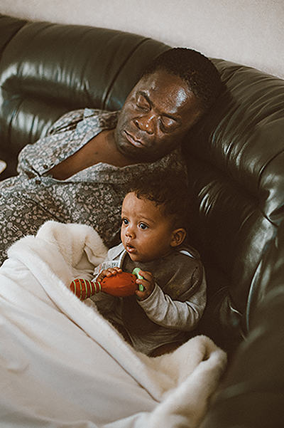 Father and toddler son sit on leather couch under a white blanket, father sleeping