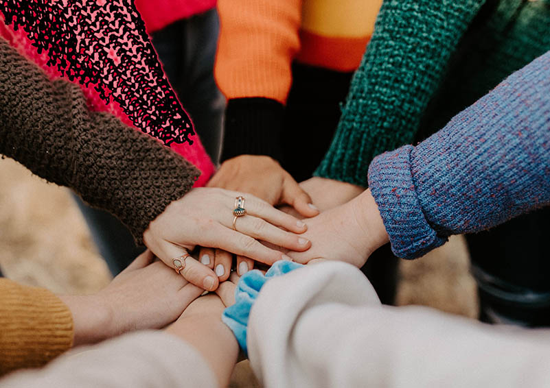 multi-racial grouping of hands, overlapping in center of a circle, arms clothed in colorful sweaters, showing cooperation