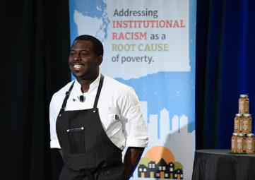 Black chef in a white shirt and black apron in front of a banner reading Addressing INSTITUTIONAL RACISM as a ROOT CAUSE of poverty.