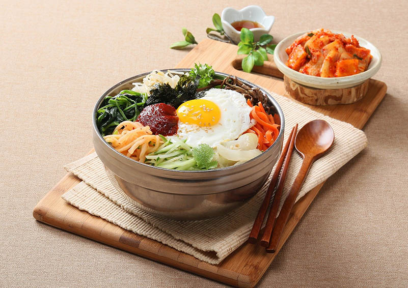Bowl of colorful veggies and a fried egg on a cutting board and napkin, with bamboo chopsticks and spoon and small bowls of condiments