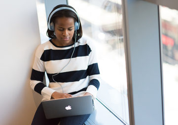 Black teen girl in black and white striped top sits in an office window working on a laptop with headphones on.