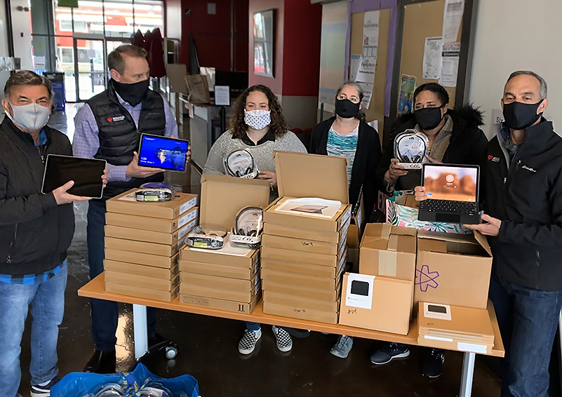 Three men and three women wearing masks pose behind a table of donated tech tools (headsets, tablets, etc.)