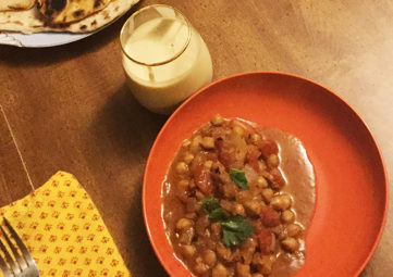 Table setting with a bright orange plate with Butter Chickpeas on it, a glass of kefir, the corner of a plate of naan, and a bright yellow napkin with a fork on it