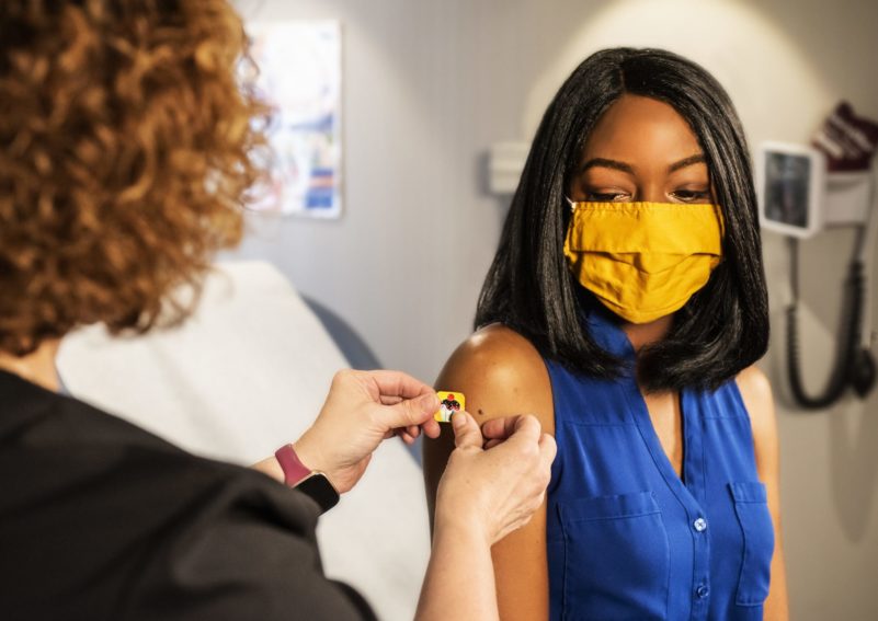 A Black woman wearing a blue sleeveless top and a mustard yellow facemask gets a bandaid from a nurse on her arm over her vaccination site