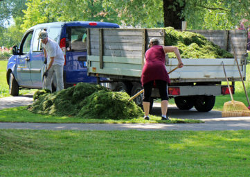 Two landscape gardeners rake a big pile of grass next to a blue van with a trailer on it.