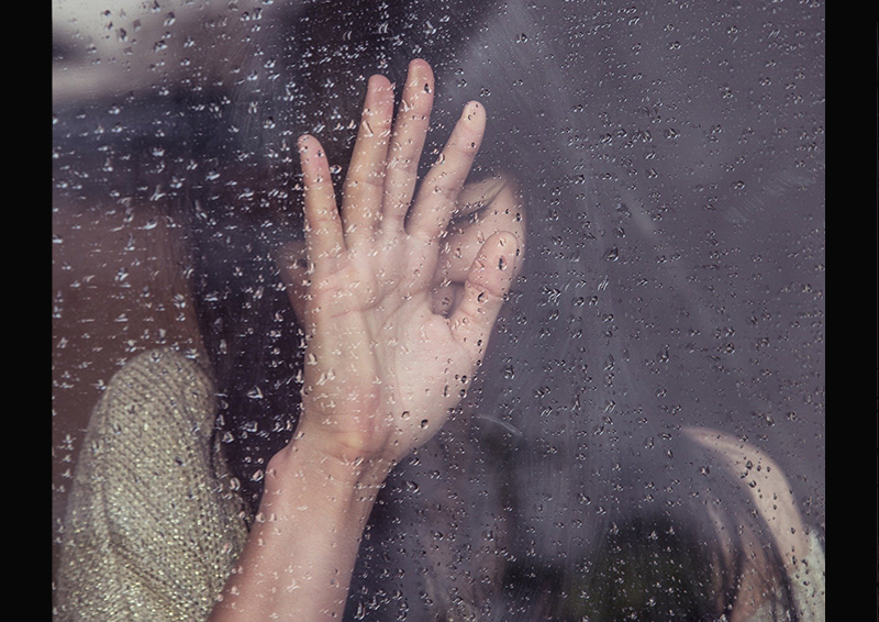 Asian woman with long black hair and a beige top leans into her hand against a rain-streaked window