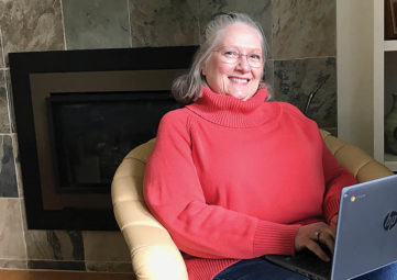 White woman with grey hair and wire-rimmed glasses, wearing a red turtleneck sweater, sitting in a beige chair with a laptop on her lap
