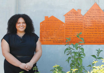 Black woman with curly shoulder-length hair wearing all black stands in front of a concrete wall with a commemorative orange plaque in the shape of houses, listing donor names.