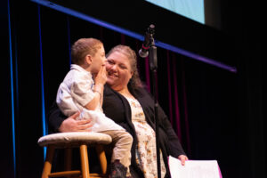 A woman on a stage smiles as she puts her arm around a small boy seated on a stool.