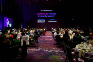 Crowded tables in a darkened ballroom, with the words "Building pathways beyond poverty" projected in blue and purple on the wall behind them.