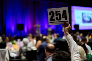 A bid card with the number 254 is thrust in the air in a convention center ballroom. Everything else is out of focus.