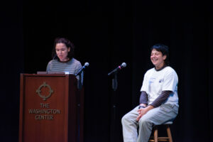 A person seated on a stool laughs as a woman stands at nearby lectern that reads "The Washington Center".