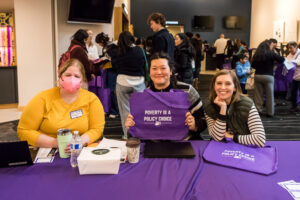 Three smiling people sit at a table in a crowded room, one of them holding up a purple cloth bag that says "POVERTY IS A POLICY CHOICE".
