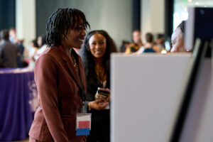 A woman in a brown suit stands next to a posterboard on an easel and talks with others.