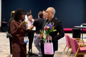A woman in a brown suit talks with a man in black suit holding flowers, a gift bag, and a glass of white wine.