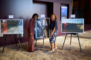 Two women pose next to a large dynamic black-and-white photo of a musician playing a saxophone.