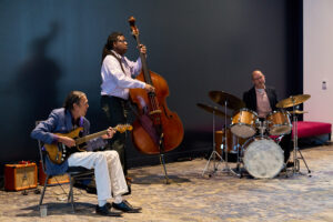 A jazz trio performs with a guitar, upright bass, and drum set.