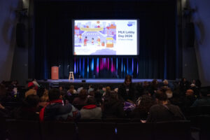 A crowd of people in a dark room facing an empty stage in front of a screen that says "Dia Legislativo de 2026" next to a cartoon illustration of people at a city intersection.