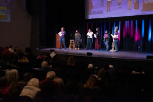Six people stand on a state, one at a podium, in front of a large audience in a dark room.